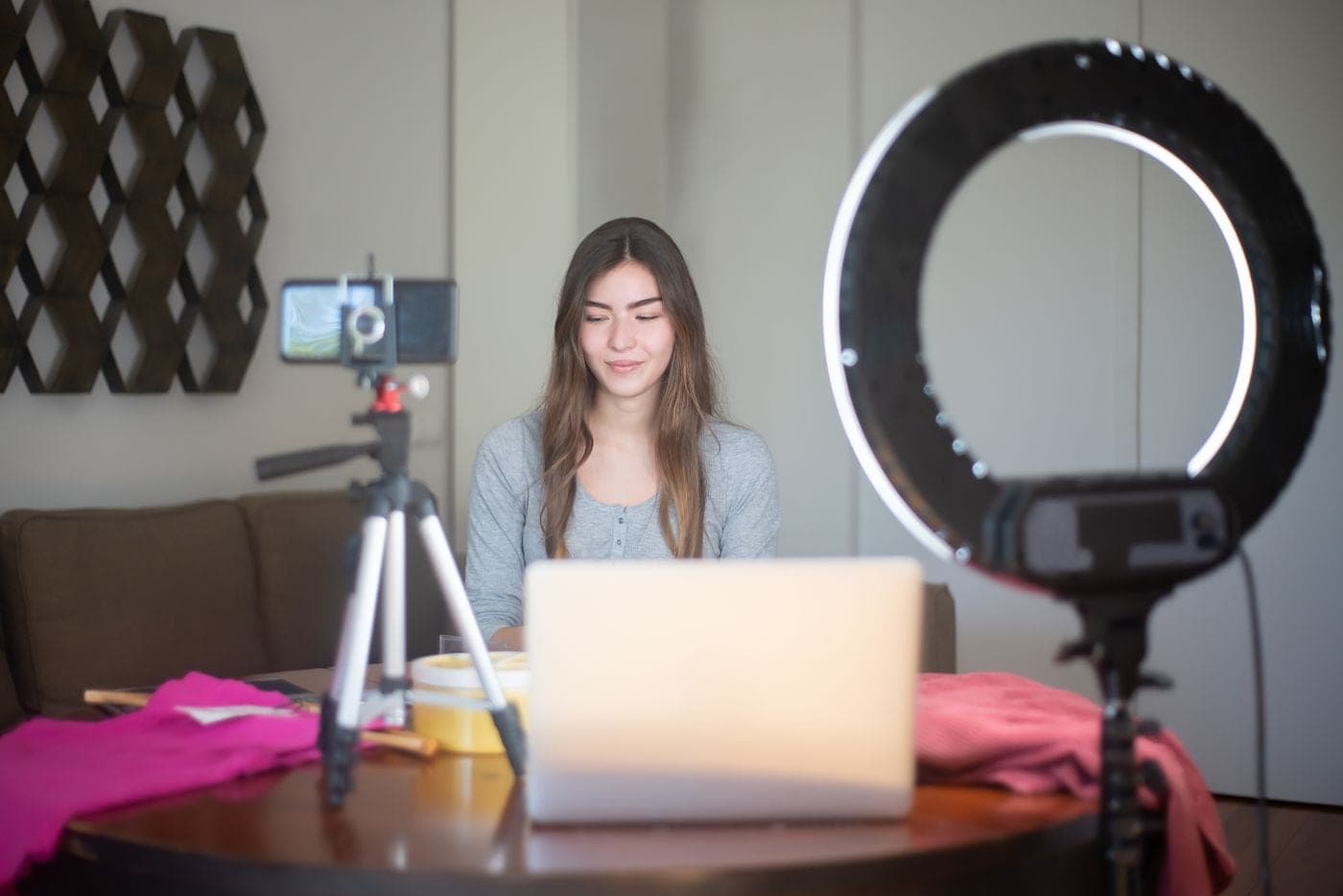 Creator vlogging indoors with a smartphone on a tripod, ring light and laptop in a home studio setup.