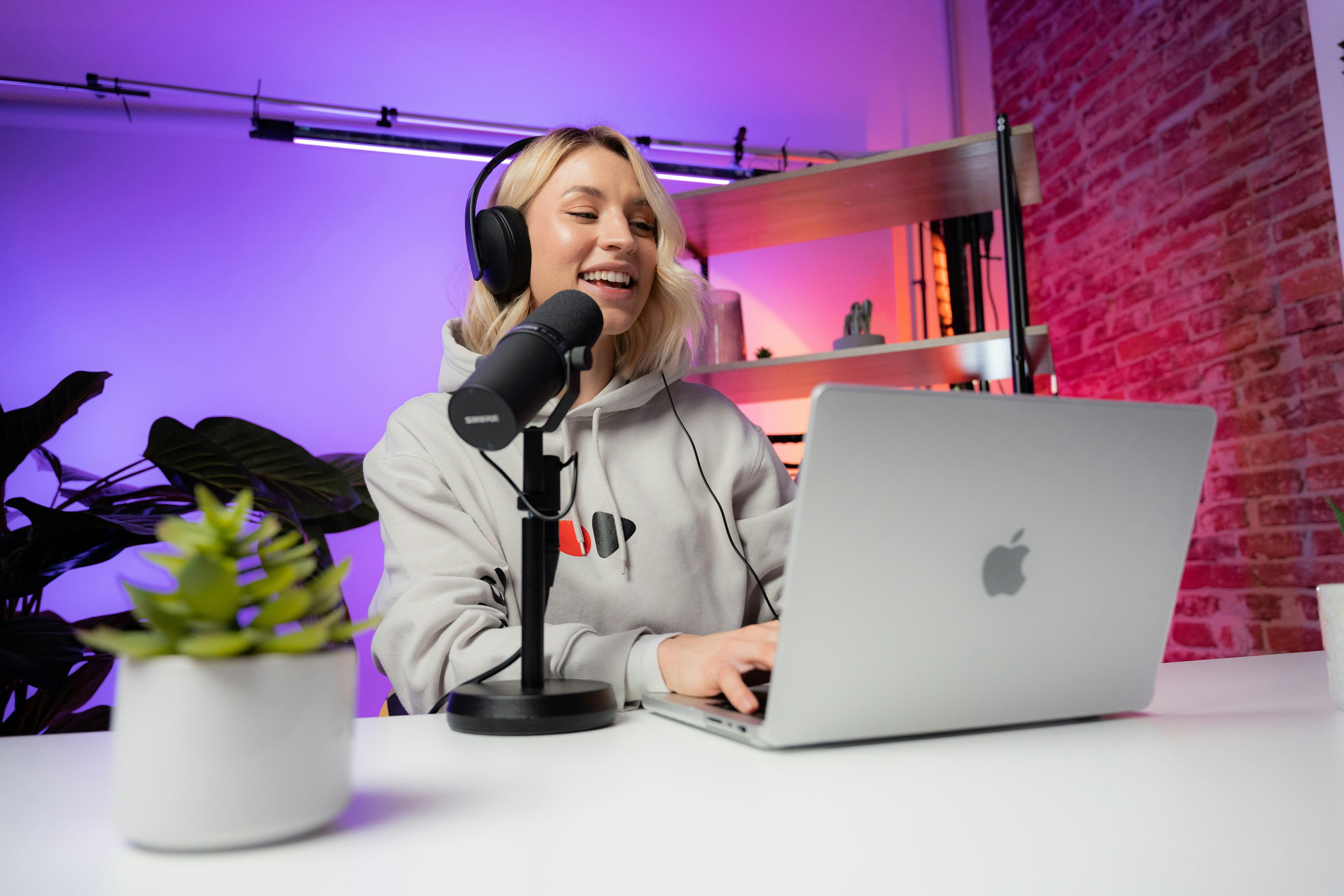 A content creator wearing headphones at a podcast studio desk with a broadcast microphone, laptop and vibrant purple and blue studio lighting.
