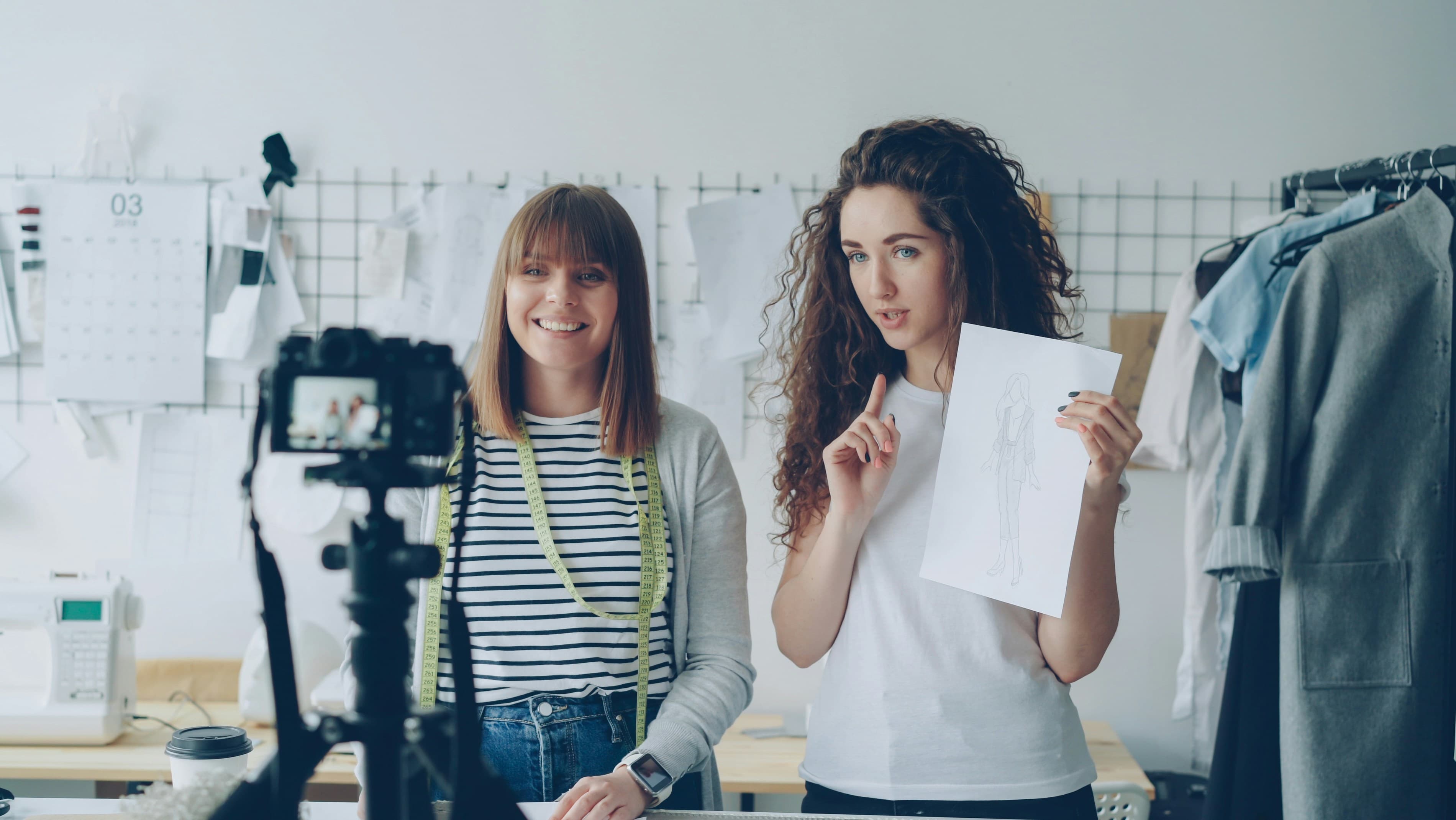 Two young content creators filming together in a bright studio with a camera, sketches and production gear visible as they create social media content.